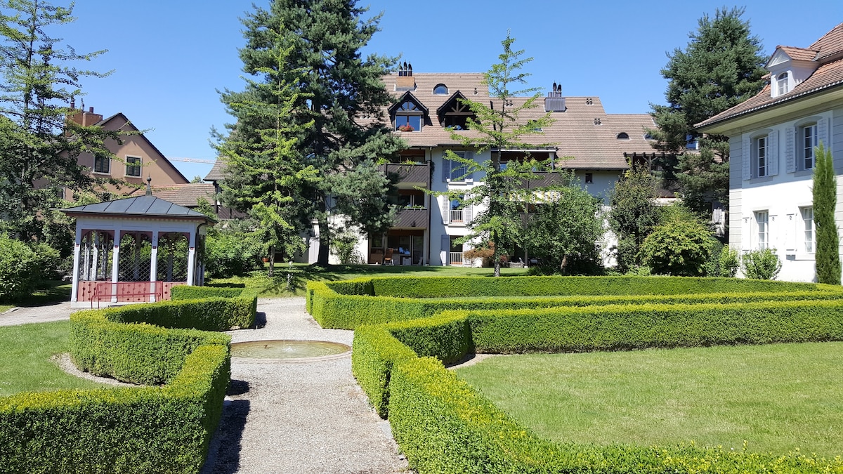 A well-maintained garden area features manicured hedges and a central fountain. Surrounding buildings, designed in a traditional style, are complemented by mature trees. A gazebo is positioned at the edge of the garden, providing a serene spot within this landscaped green space.