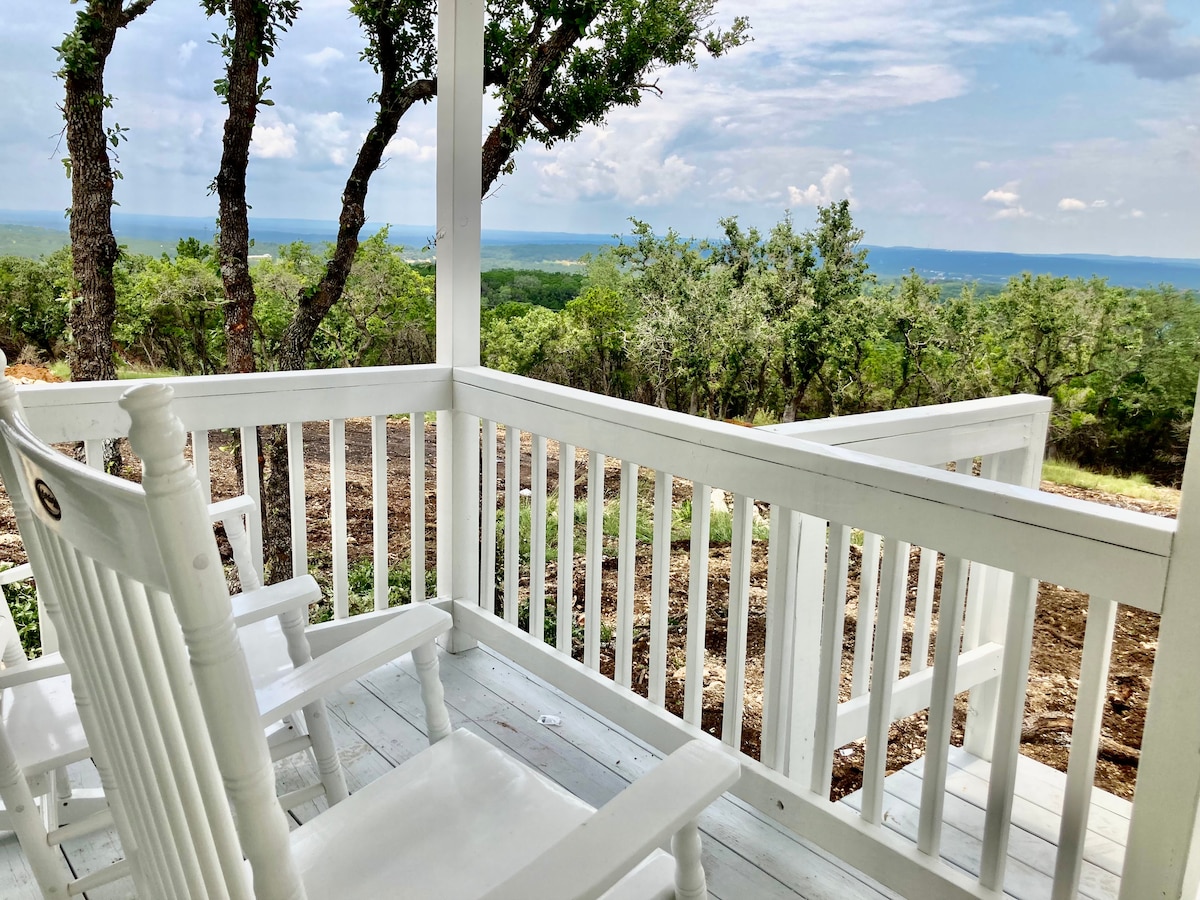 A spacious porch features white rocking chairs, offering relaxing seating. The surrounding trees and rolling hills expand into the horizon, with a backdrop of cloudy skies. Nature’s beauty is highlighted, creating a serene outdoor space for enjoying the view.
