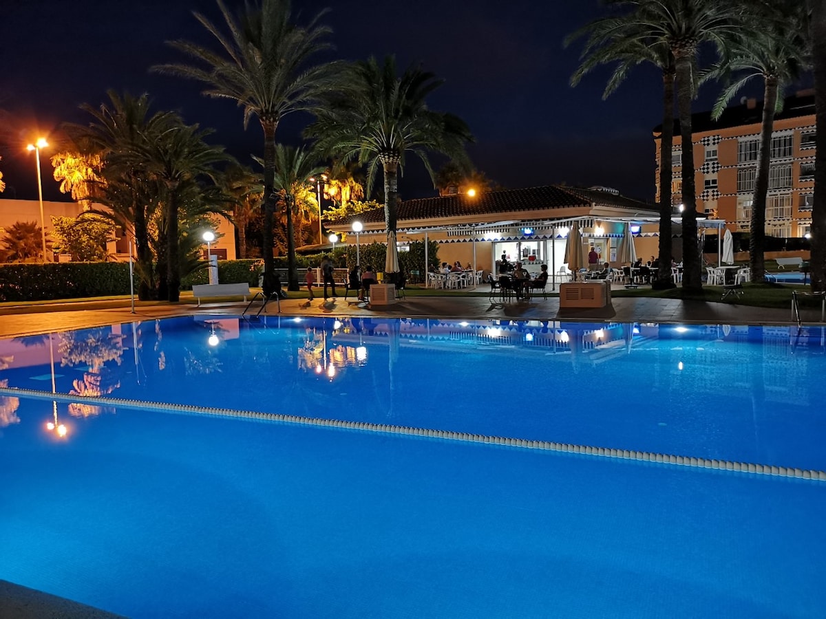 A well-lit pool area is surrounded by palm trees, creating a relaxing ambiance at night. The water reflects the surrounding lights, enhancing the serene atmosphere. In the background, a building with outdoor seating areas is visible, contributing to the inviting environment.