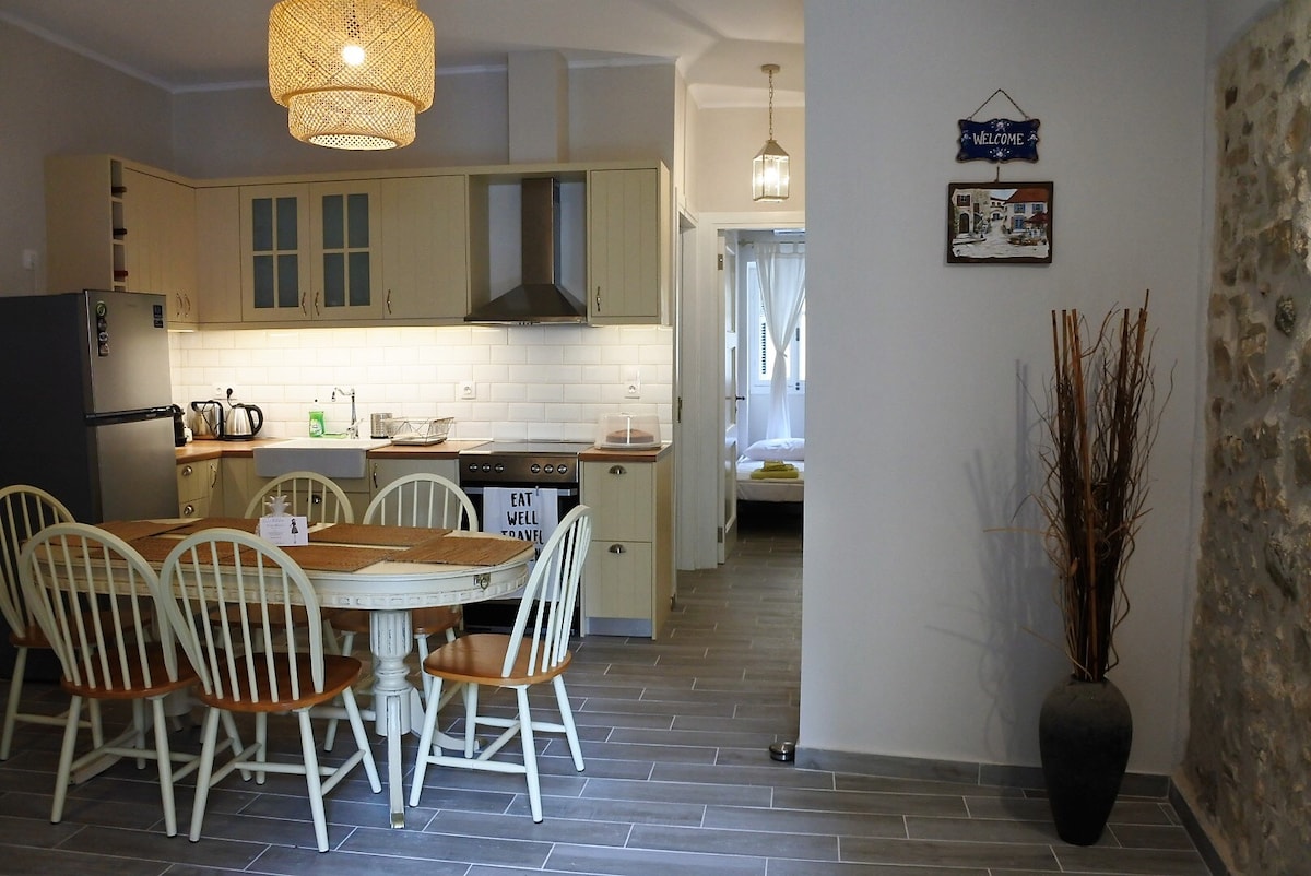 A fully equipped kitchen is visible, featuring light-colored cabinetry and a gray tiled floor. A round wooden dining table with six chairs is positioned at the center. A stone wall adds texture to the space, complemented by decorative elements and an open layout leading to another room.