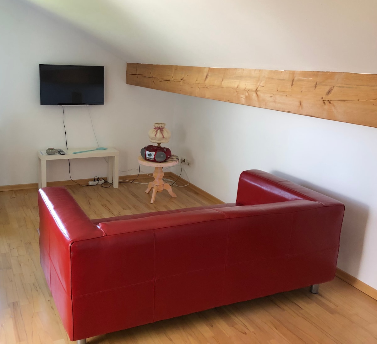A seating area features a modern red couch positioned against a wall, complemented by a small side table holding a decorative item. A flat-screen TV is mounted on the wall, and natural light fills the room, highlighting the wooden accents of the space.