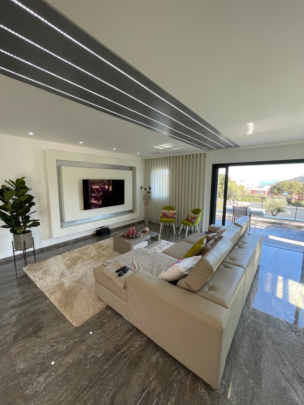 A modern living area is highlighted by a sleek sectional sofa and a large wall-mounted television. Natural light fills the space through floor-to-ceiling windows, while a plush area rug and decorative chairs add comfort. A potted plant brings a touch of greenery to the environment.