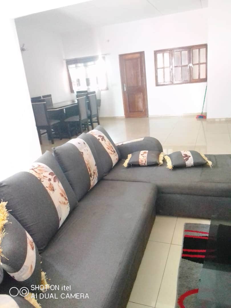 A spacious living area is featured with a dark gray sectional sofa, adorned with decorative cushions. The floor is tiled, and a patterned rug is placed in front of the sofa. In the background, a dining area with a dark table and chairs is visible through seated windows.