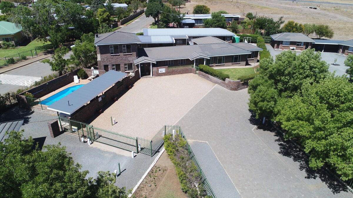 An aerial view of Sunset Chalets showcases the layout of the accommodation, featuring a secure entrance with a large parking area. A tranquil blue pool is visible, bordered by shaded areas, with spacious gardens and pathways leading through the property.