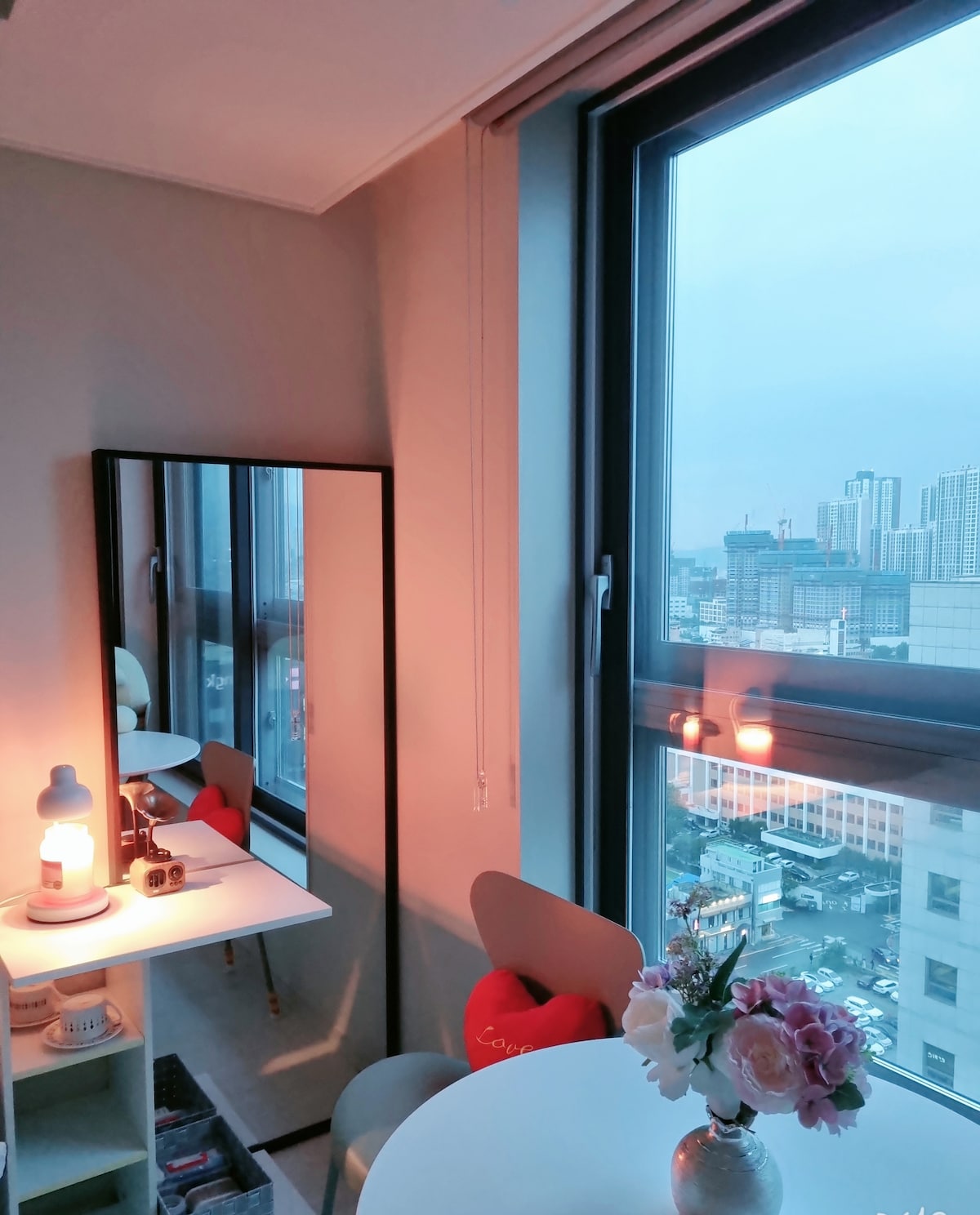 A well-lit corner features a mirror and a small table with decorative items. A vase with flowers adds a touch of color. Large windows offer a view of the city skyline, with soft lighting creating a calming atmosphere.