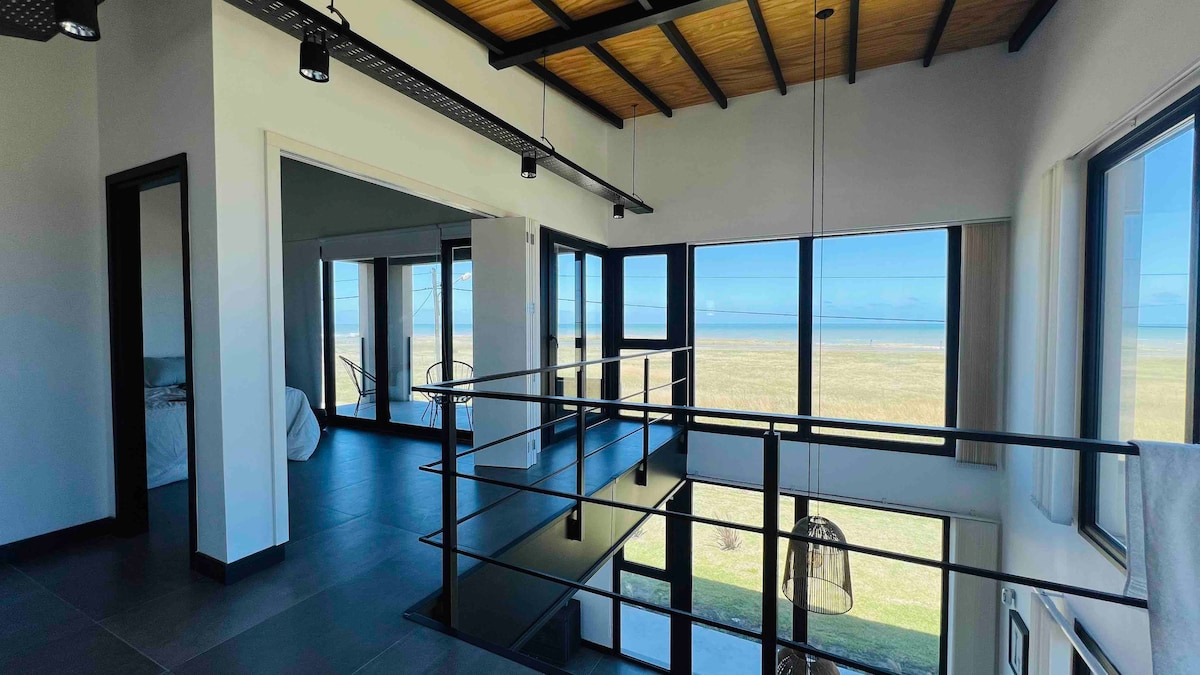 A spacious room with large windows offers panoramic views of the sea and surrounding landscape. The contemporary design is enhanced by wood-beamed ceilings and a sleek railing along the upper level. Abundant natural light floods the space, creating an open and airy atmosphere.