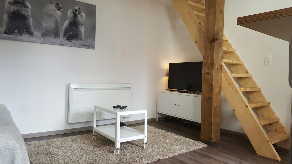 The living area features a neutral color scheme with a cozy rug under a simple coffee table. A television is mounted on a white media console. Wooden beams and a staircase contribute to an airy ambiance. A black-and-white artwork of animals adorns the wall.