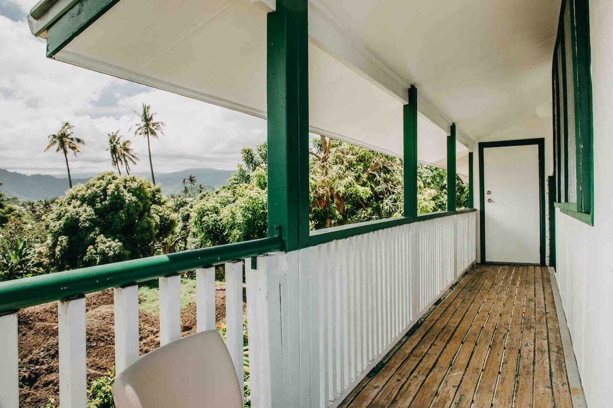 An open balcony space features wood and white railings, surrounded by lush greenery. Nearby, tall palm trees are visible against a cloudy sky. A door leads to an interior space, while sunlight highlights the natural wood flooring.