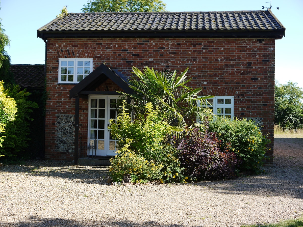 The exterior of Rose Cottage is presented, featuring warm red brick and traditional windows. Lush greenery, including a palm and flowering shrubs, frames the entrance, welcoming visitors. A gravel pathway leads to the front door, enhancing the cottage's rustic charm.