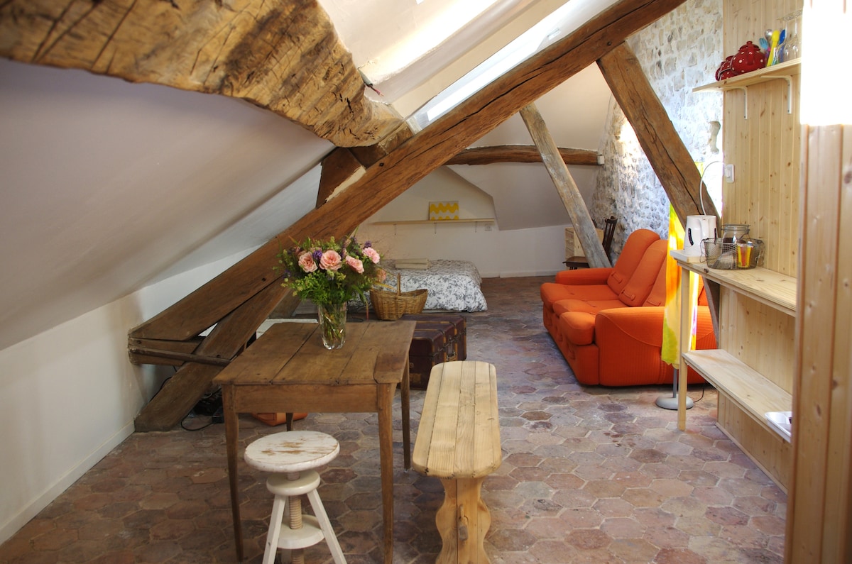 A charming attic space features exposed wooden beams and rustic stone walls. A cozy seating area with an orange sofa faces a simple wooden table and two benches, while a spacious bed is positioned against the wall. Natural light brightens the room, enhancing its inviting character.