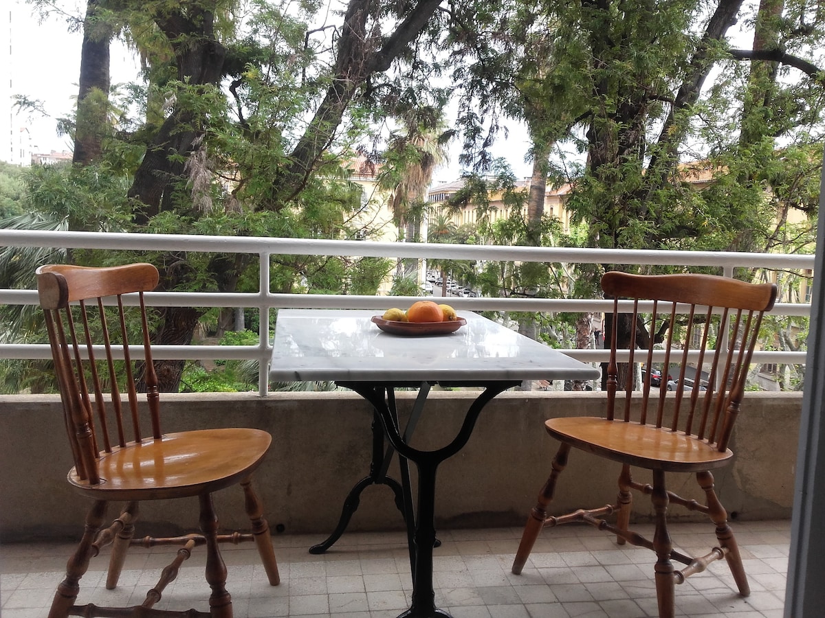 A small balcony features a white table with a bowl of fruit resting on it. Two wooden chairs with curved backs are positioned around the table. Lush greenery is visible beyond the railing, providing a calming view.