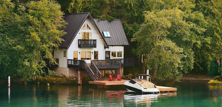 Une photo montre un bateau, couvert et amarré devant une maison de 2 étages située au bord d'un lac.