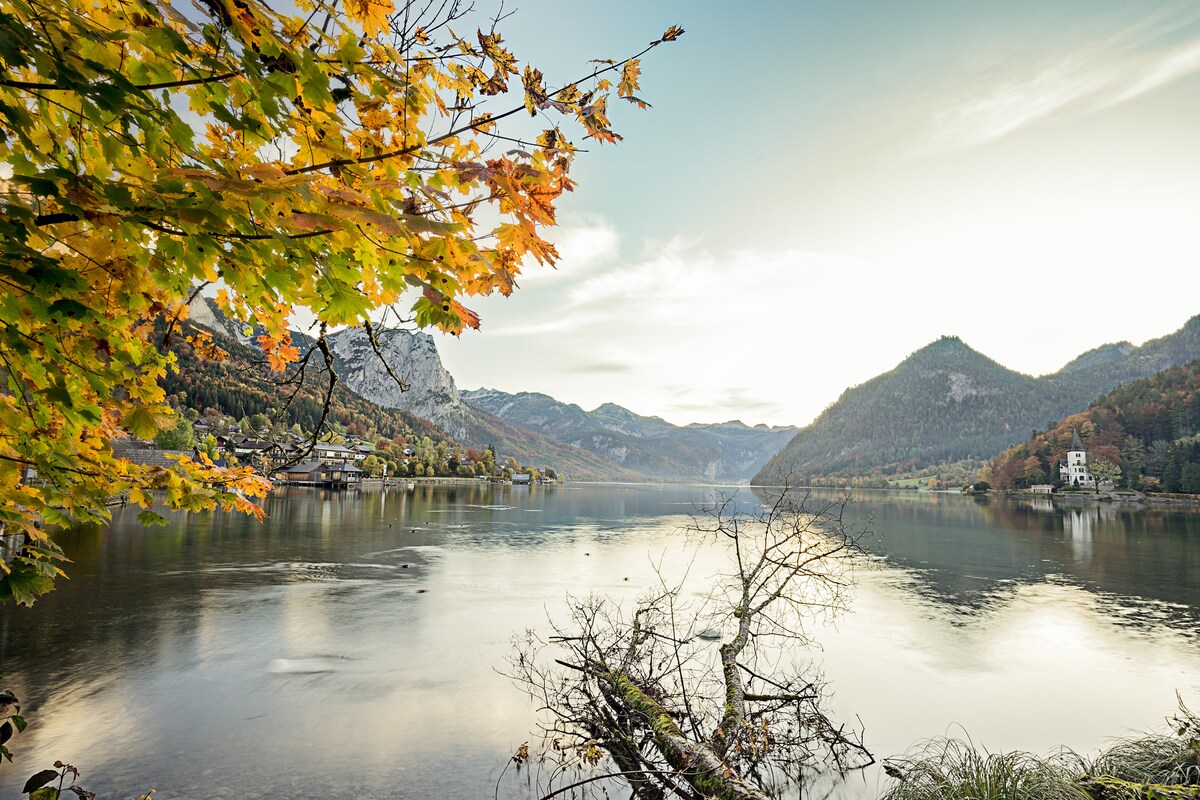 A serene lake is framed by vibrant autumn foliage and majestic mountains in the background. The calm water reflects the soft light of the sky, creating a tranquil atmosphere. The scene highlights the natural beauty of the surroundings amidst the changing seasons.