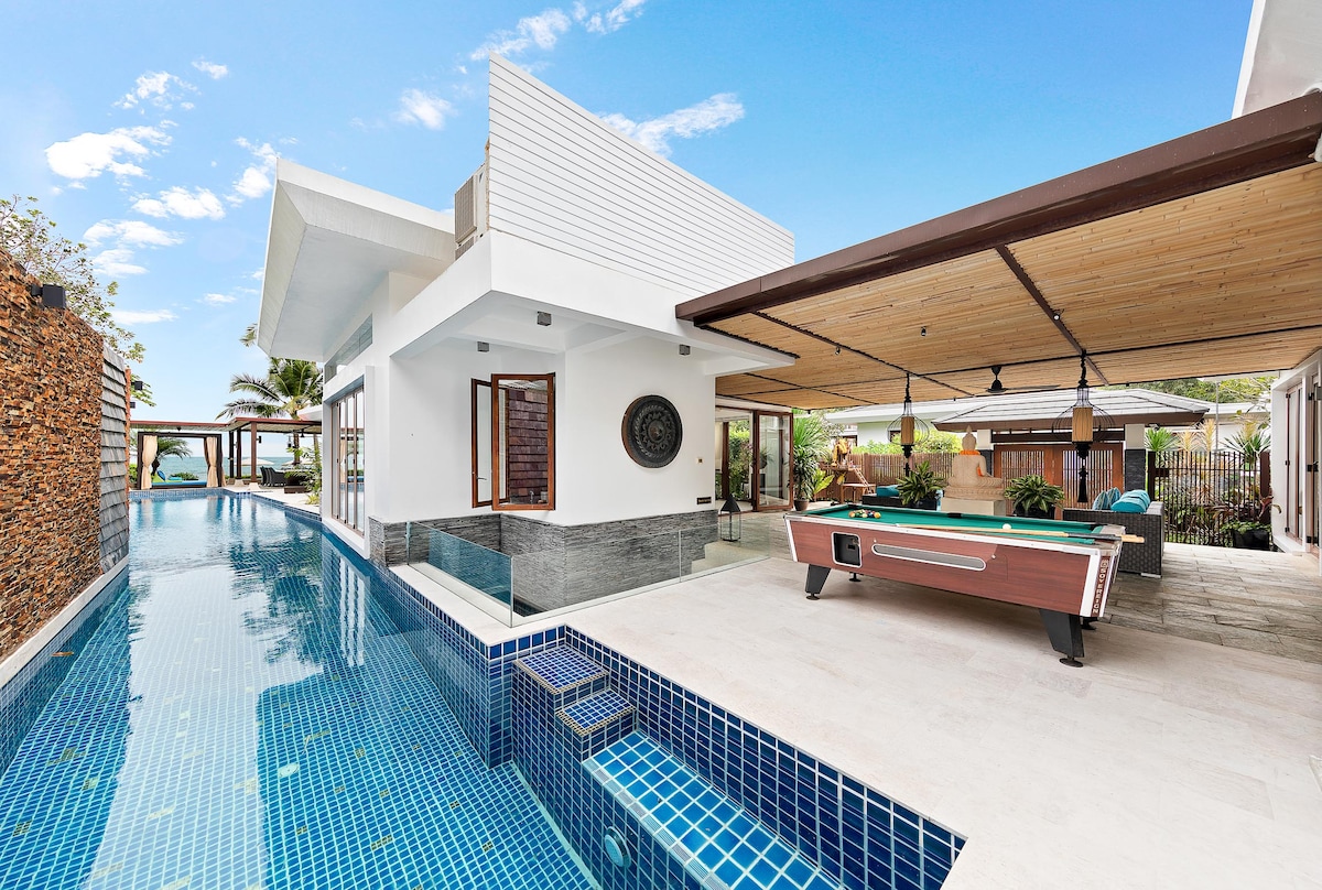 A modern villa features a private lap pool alongside a spacious outdoor area. A billiards table is positioned beneath a covered terrace, while large windows provide views of the surrounding landscape and water. Tropical plants enhance the inviting atmosphere.