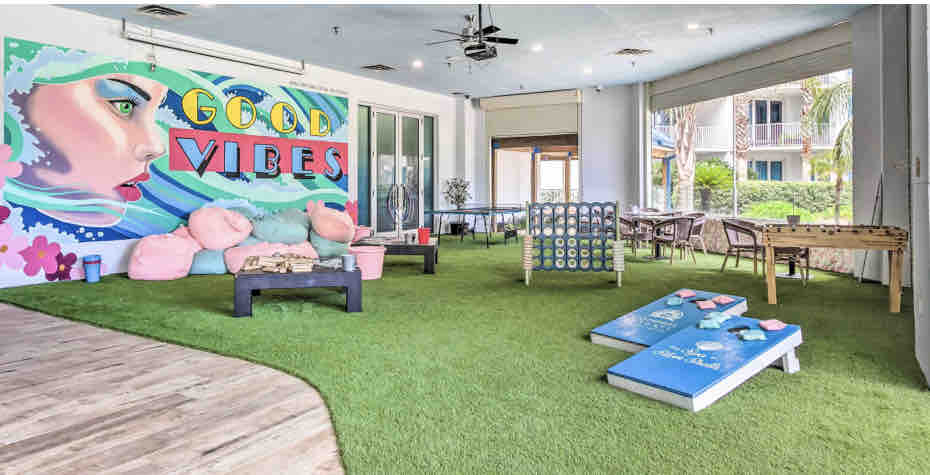 A communal lounge area features artificial grass flooring with seating options including colorful bean bags and a low table. A vibrant mural stating 'GOOD VIBES' adorns the wall, while nearby activities include a foosball table and a game area designed for leisurely play.