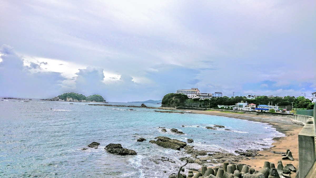 A scenic view of a tranquil beach is presented, with gentle waves lapping against the shore. Coastal rocks are visible along the water's edge, while a cluster of buildings sits in the distance, surrounded by lush greenery under a cloudy sky.