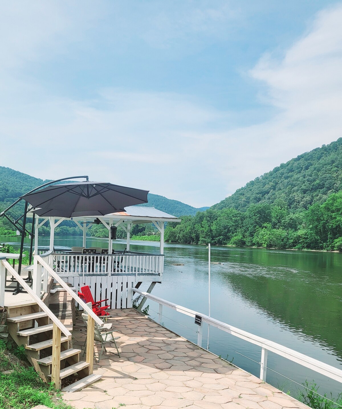 A shaded outdoor seating area overlooks the New River, with a protective umbrella casting shade over a chair. The patio features a wooden railing, and steps lead down to the riverbank, surrounded by lush green hills and clear blue skies in the background.