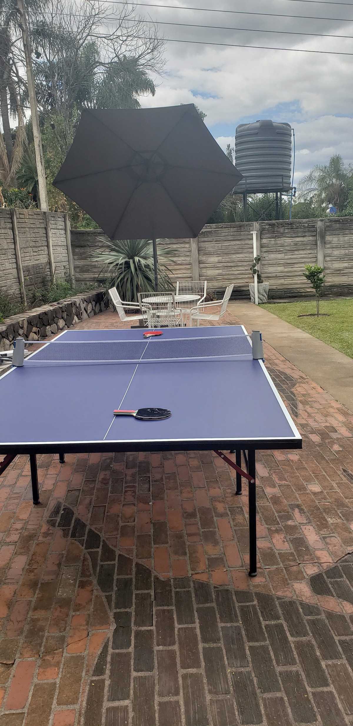 A ping pong table is set up on a brick patio, with a large umbrella providing shade. White outdoor chairs are positioned nearby. In the background, a water tank is visible, surrounded by greenery and a stone wall, creating a relaxed outdoor setting.