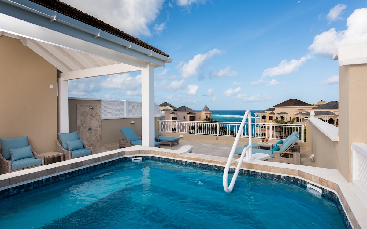 The rooftop pool is framed by a spacious patio area, featuring multiple sun loungers for relaxation. A panoramic view of the ocean is visible in the background, with soft clouds scattered across a bright blue sky.