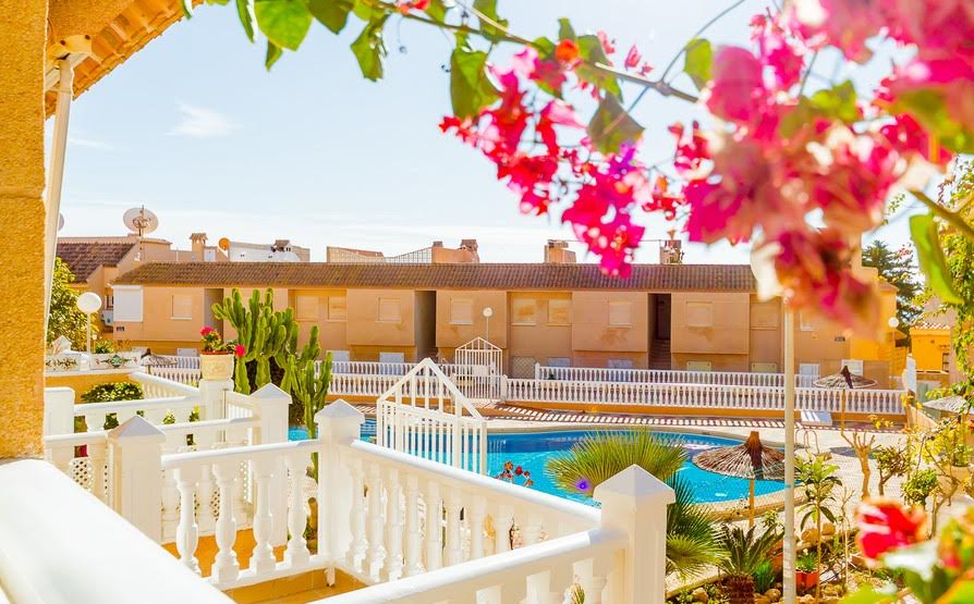 A view of the outdoor area features vibrant pink flowers framing the scene. A pool is visible in the center, surrounded by well-maintained greenery and a white fence. Several structures with beige exteriors are seen in the background under a clear sky.