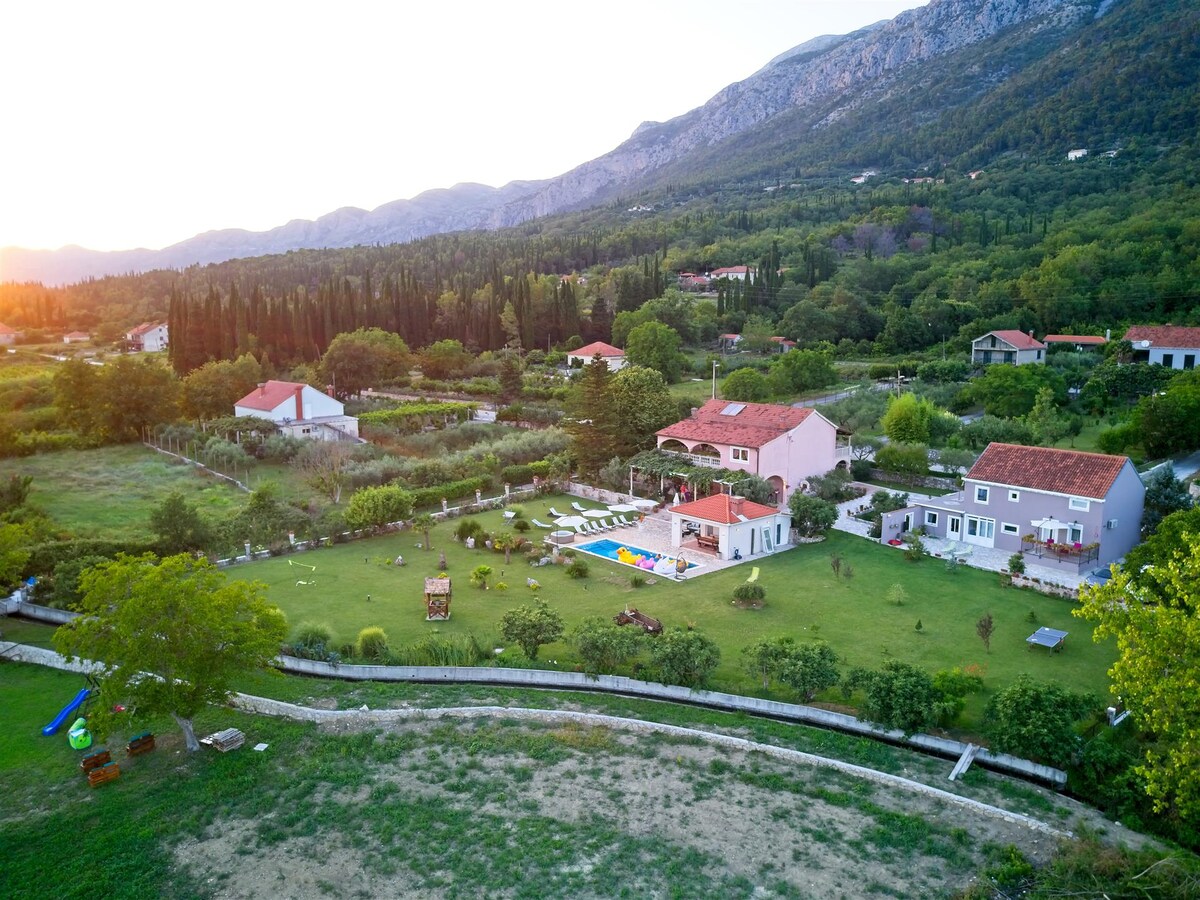 An aerial view of Villa Thousand Roses is displayed, surrounded by lush greenery and mountains. The property features a spacious garden with a private pool and several buildings, including the villa and nearby cottages, amidst the vibrant landscape.