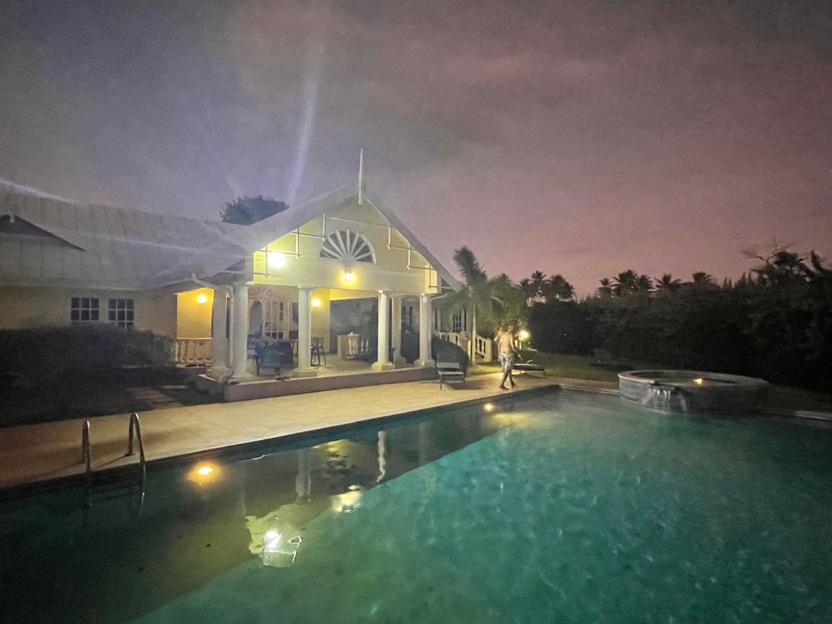 The exterior of a villa is illuminated at night, featuring a spacious pool and a porch area. The building reflects a blend of architectural styles with large windows and a balcony. Lush greenery surrounds the property, enhancing the serene atmosphere.
