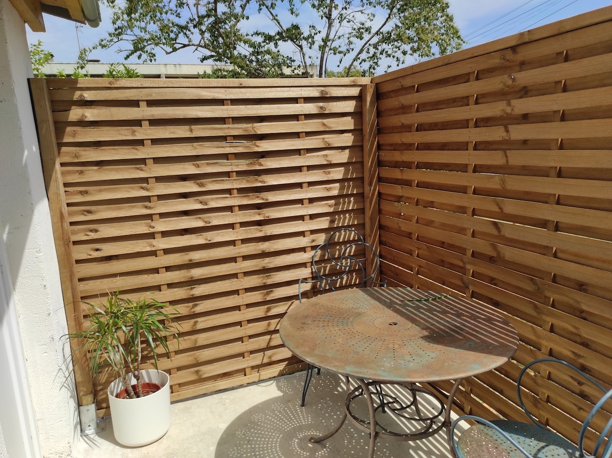 A small terrace is enclosed by wooden slats, providing privacy. A round metal table is set alongside two decorative chairs. A potted plant adds a touch of greenery, and sunlight casts decorative shadows on the ground.