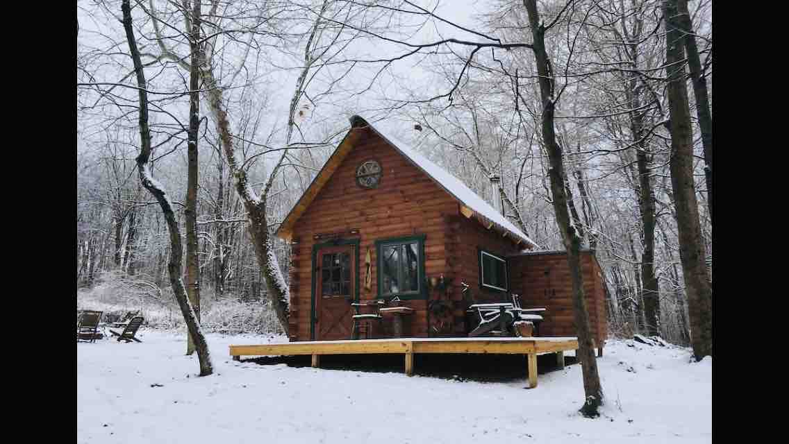 A cabin constructed of wood is surrounded by snow-covered trees. A small deck extends from the front, where outdoor seating is arranged. A large window allows for views of the snow-blanketed landscape. The cabin’s rustic charm is enhanced by its natural setting.