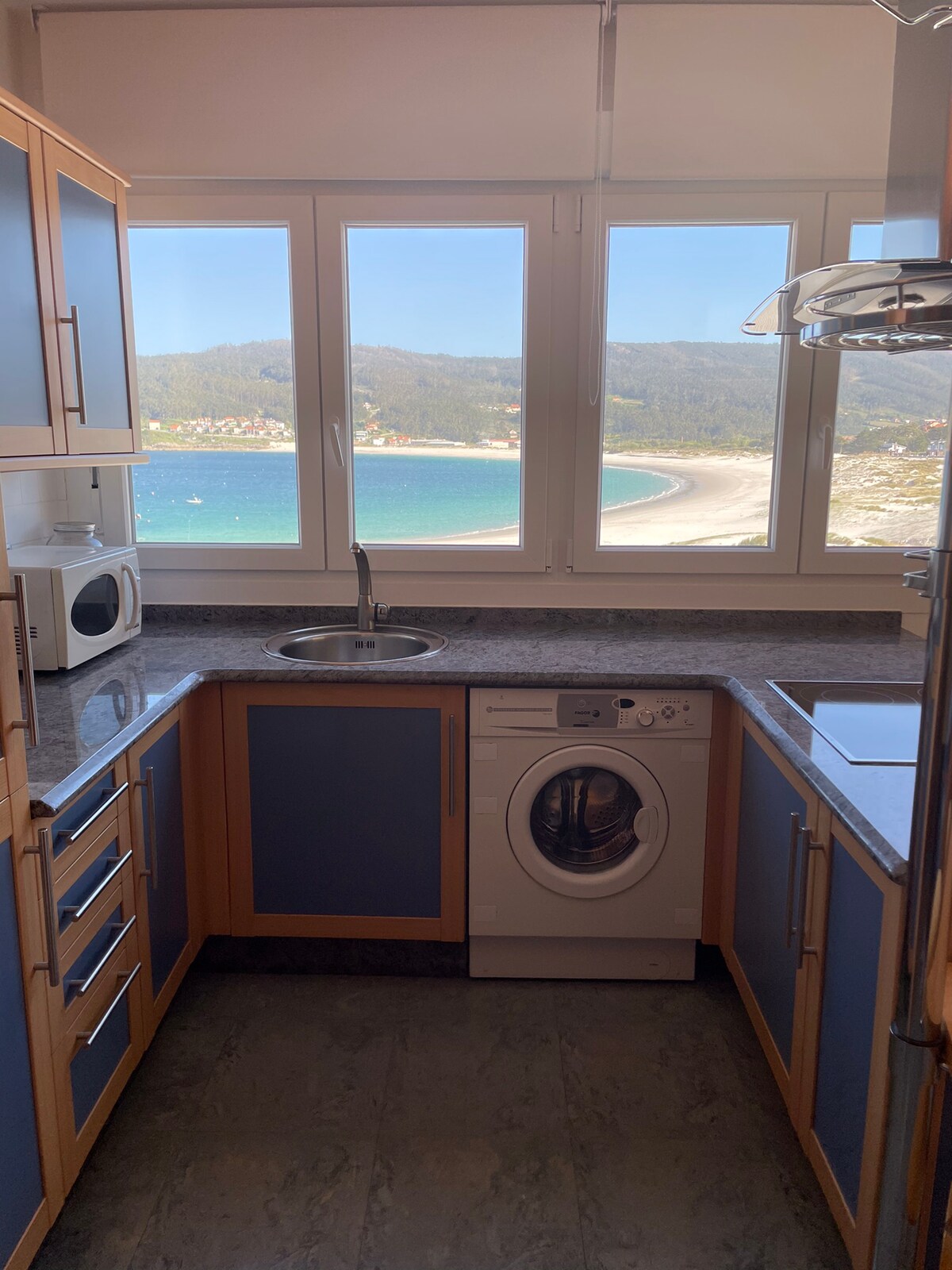 A well-equipped kitchen features modern appliances including a microwave and a washing machine. The countertop is complemented by light wood cabinetry, while large windows showcase scenic views of the beach and coastline.