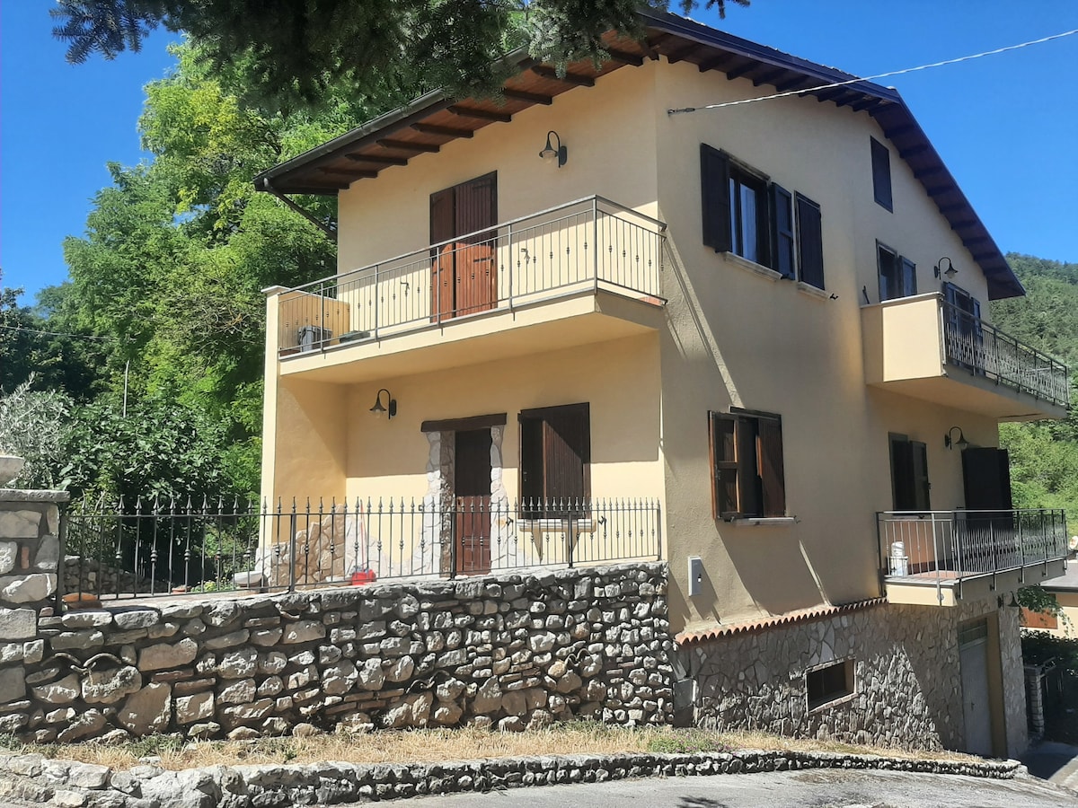A three-story house features a light-colored exterior with wooden doors and windows. The property is surrounded by a stone wall and includes balconies on the upper floors. Lush greenery and trees provide a natural setting, enhancing the peaceful environment.