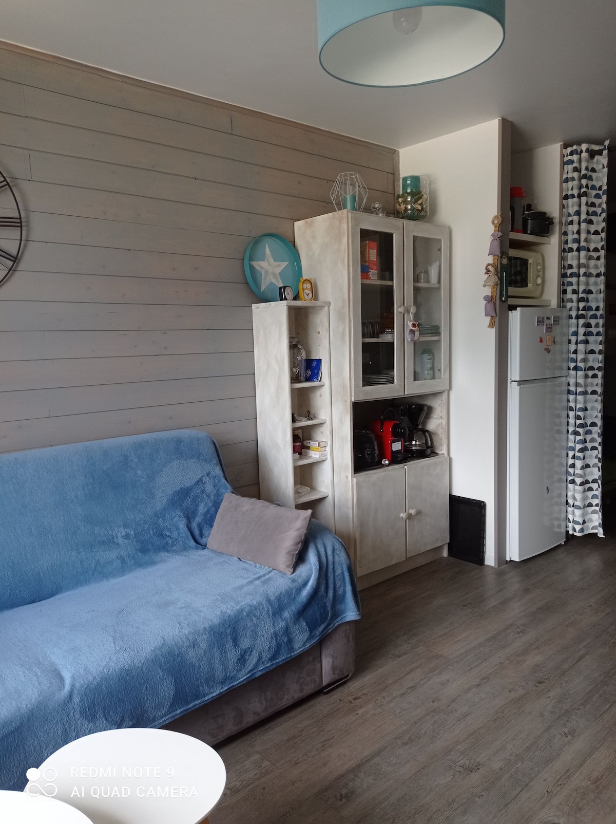 A cozy corner of the studio is highlighted by a sofa covered with a blue throw. A tall cabinet displays an assortment of items. A compact refrigerator stands nearby, while soft-colored wooden panels add warmth to the space. Natural light fills the area.
