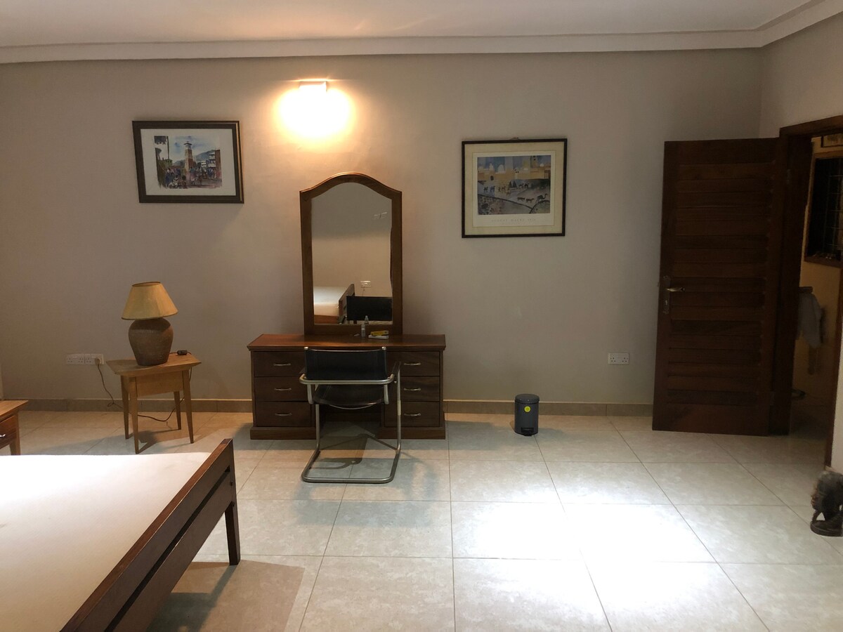 A private bedroom is showcased with a spacious layout, featuring a wooden bed frame, a dresser with a mirror, and a chair. Natural light is introduced through a window, and artwork adorns the walls. A lamp and a heater are visible, contributing to the room's functionality.