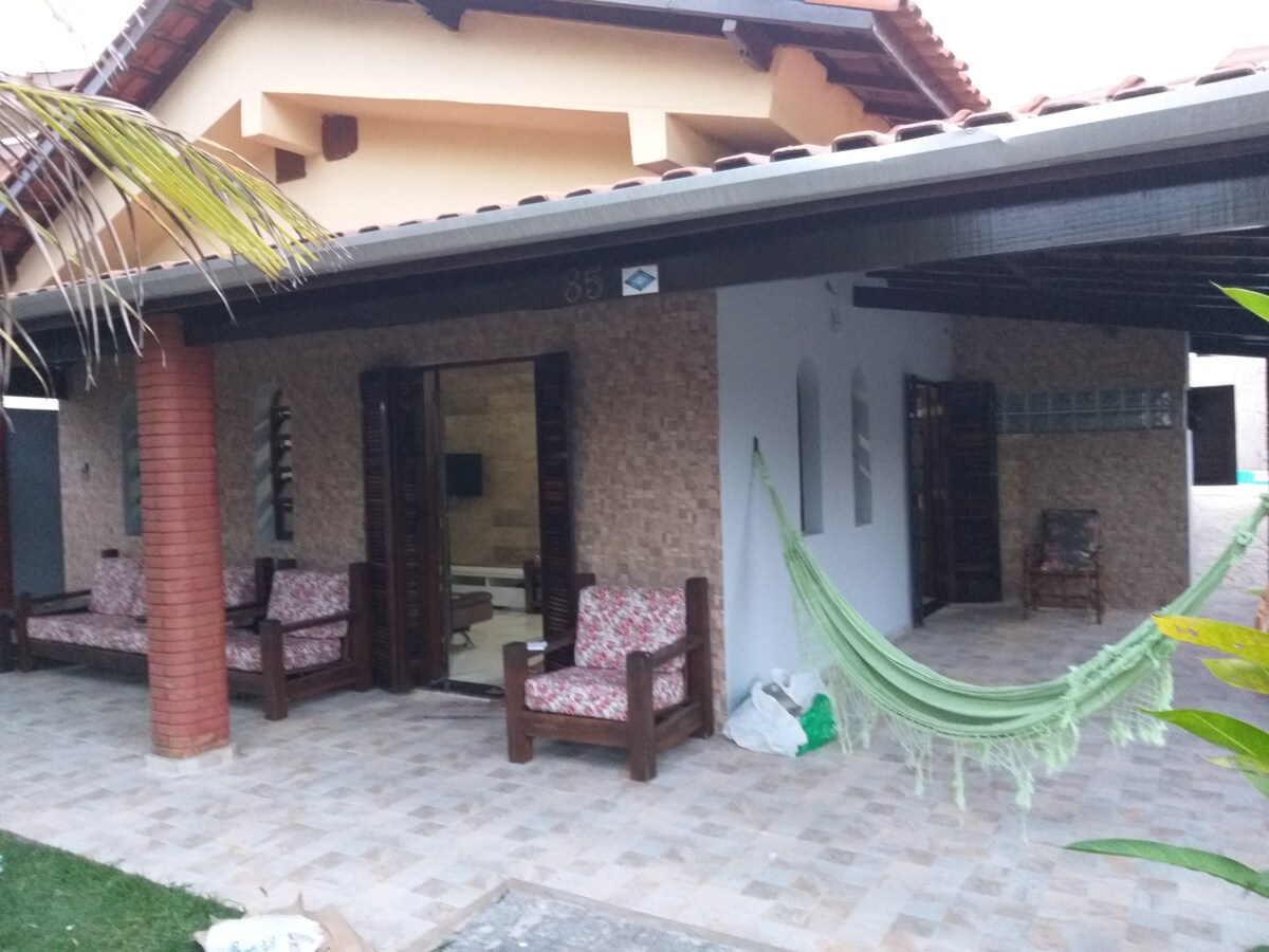 An outdoor seating area features wooden chairs with floral upholstery, inviting relaxation. A green hammock is suspended between columns, providing a comfortable spot to rest. The stone-paved patio is framed by lush greenery, and large glass doors lead into the interior of the home.