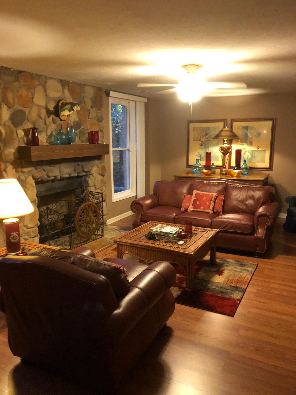 A cozy living room features a stone fireplace with an ornate grate, complemented by two leather sofas and an armchair. A wooden coffee table sits in the center, decorated with books and a small table lamp. Soft light from a ceiling fan illuminates the warm wood flooring.