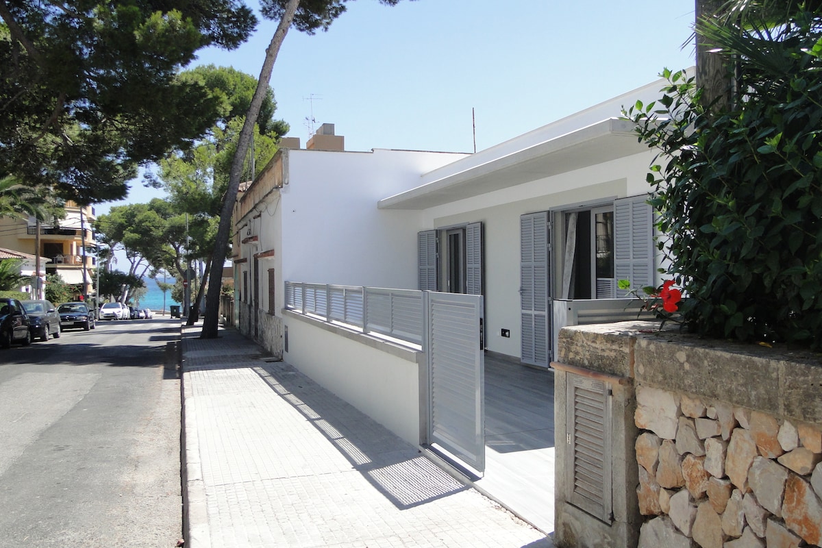A modern house exterior is shown, featuring a light façade and gray shutters. A gated entrance leads to a spacious walkway lined with smooth tiles. Surrounding trees provide shade, and distant views of the sea are visible, indicating proximity to the beach.