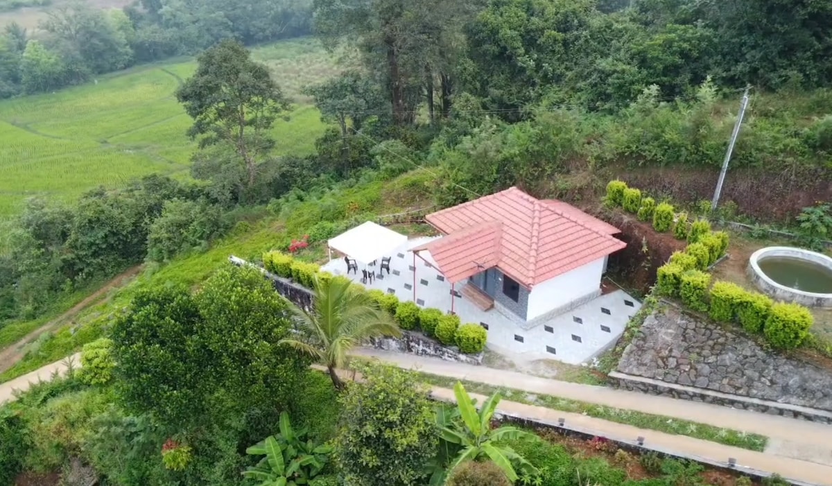 A serene aerial view of a standalone house is surrounded by lush greenery and fields. The structure features a red tiled roof and is bordered by neatly trimmed hedges. A shaded outdoor seating area is positioned nearby, enhancing the home’s tranquil setting in the countryside.