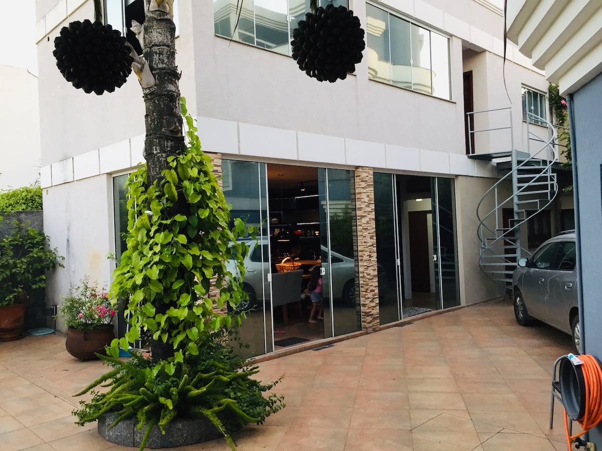 The exterior of the building is visible, showcasing a modern design with large glass windows. A spiral staircase leads to a higher level. Lush greenery and decorative plants are present in the foreground, adding a natural touch to the courtyard area.