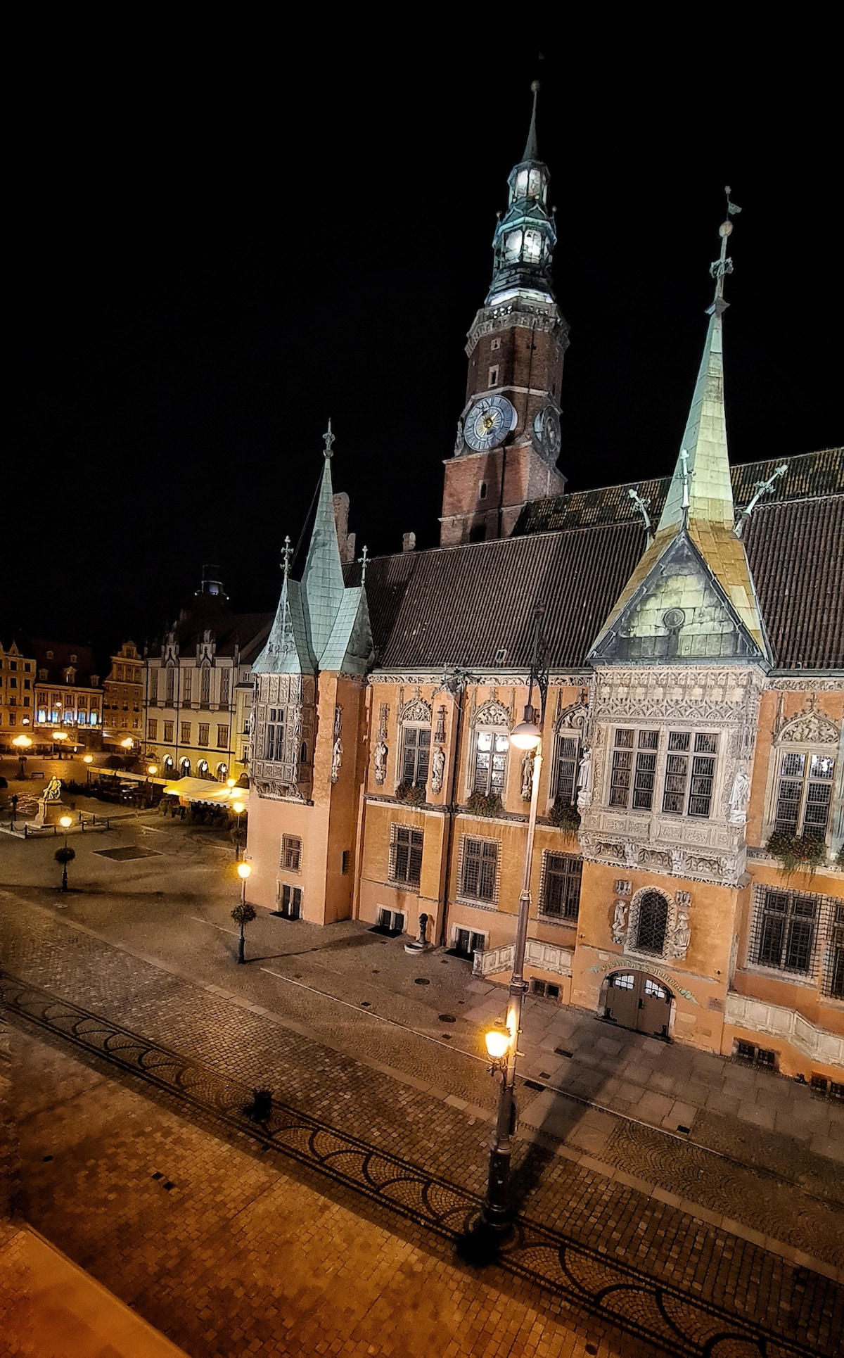 The image captures the Wrocław Town Hall illuminated at night, showcasing its distinctive architecture with pointed roofs and intricate detailing. The clock tower stands prominently, with warm street lamps casting a gentle glow on the surrounding cobblestone square, enhancing the historical ambiance of the area.