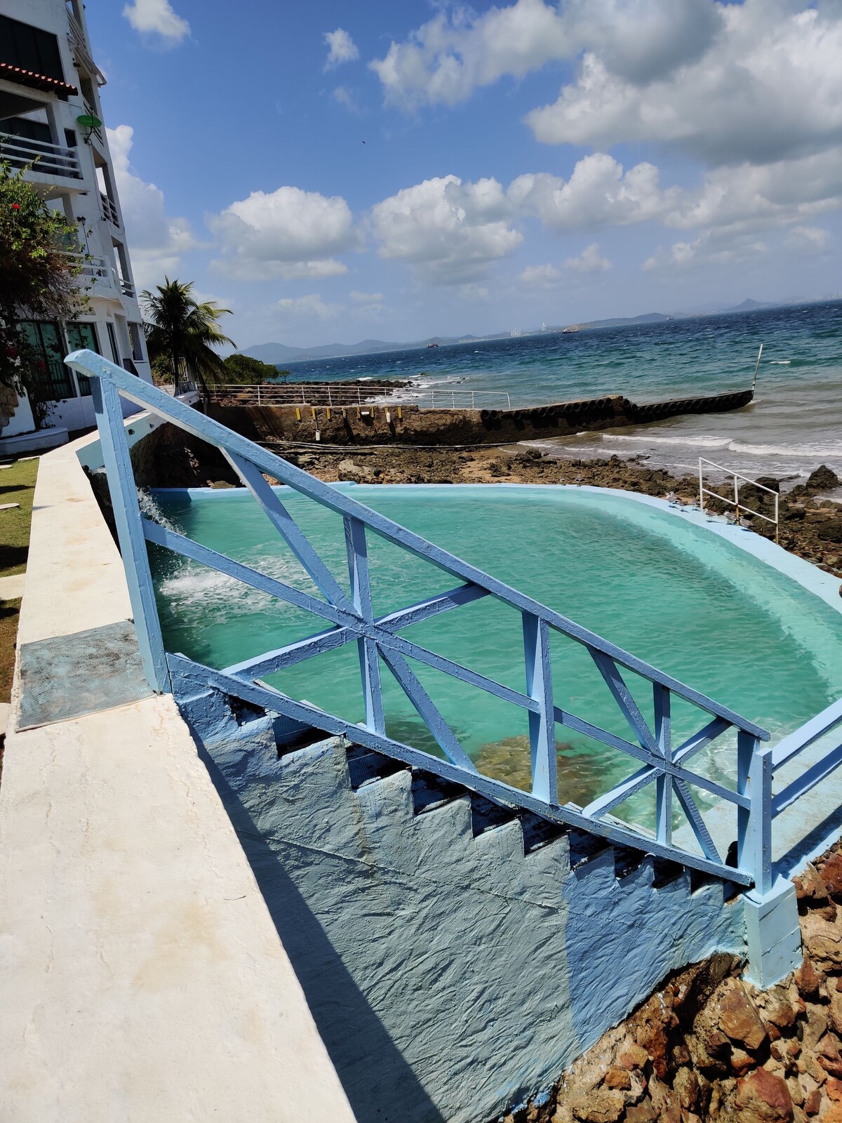 A salt-water pool is set against a coastal backdrop, with steps leading down into the water. The pool's turquoise hue contrasts with the rocky shoreline and the vibrant blue sky dotted with clouds. The calming ocean waves can be seen in the distance.