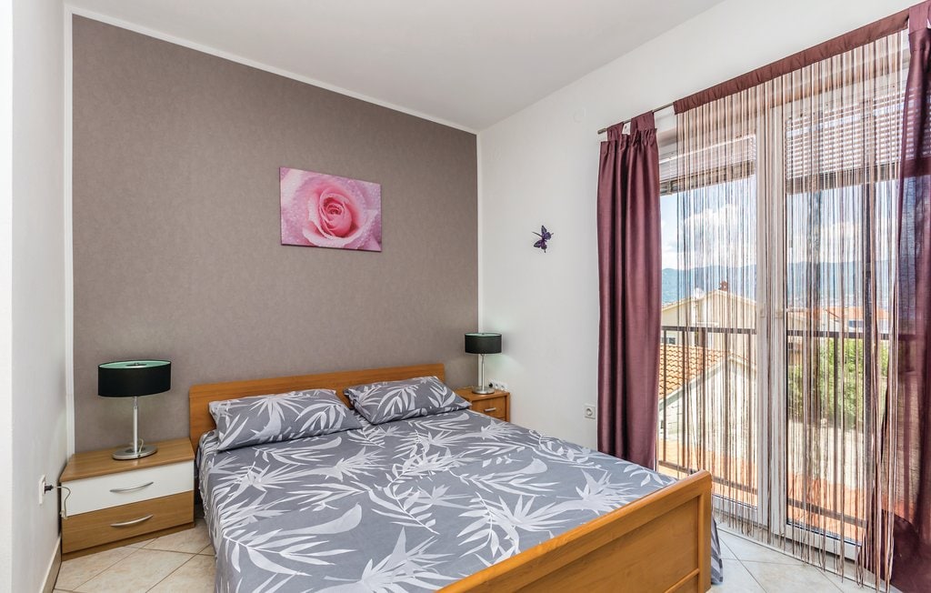 A spacious sleeping area features a wooden bed with a patterned duvet cover. Two bedside lamps provide soft lighting, and a floral wall art piece adds a touch of color. Large windows with sheer curtains allow natural light to fill the room, offering views of the surroundings.