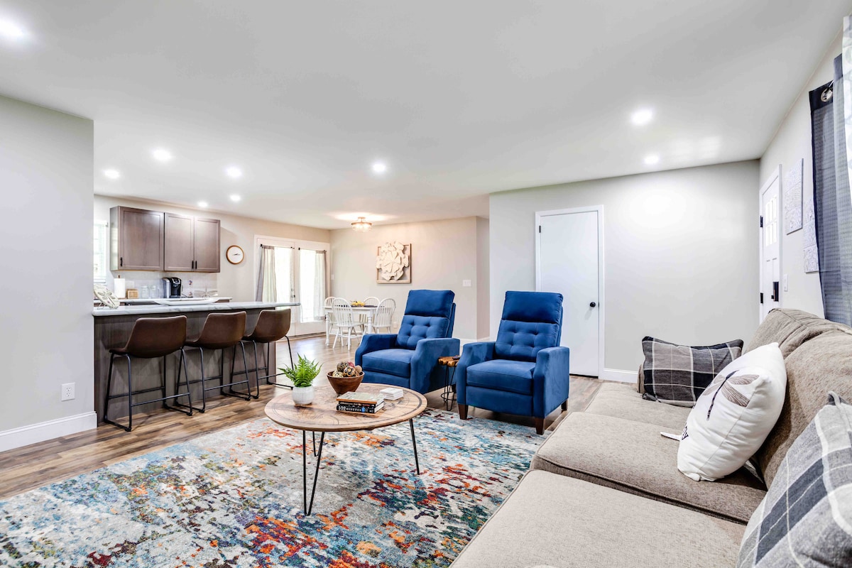 A light-filled living area is presented with a contemporary rug and a comfortable sectional sofa. Two plush blue chairs are positioned around a circular coffee table, which holds decorative items. A dining area and kitchen are visible in the background, enhancing the open layout.