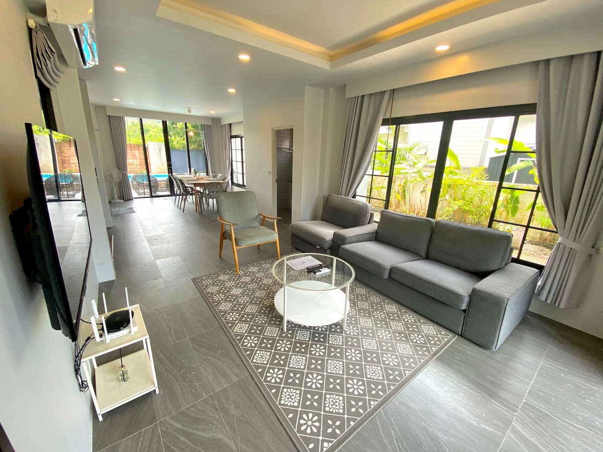 A spacious living area features large windows providing natural light, with views of lush greenery. A comfortable grey sectional sofa is arranged around a circular glass coffee table, and a modern dining area is visible in the background, accommodating multiple guests.