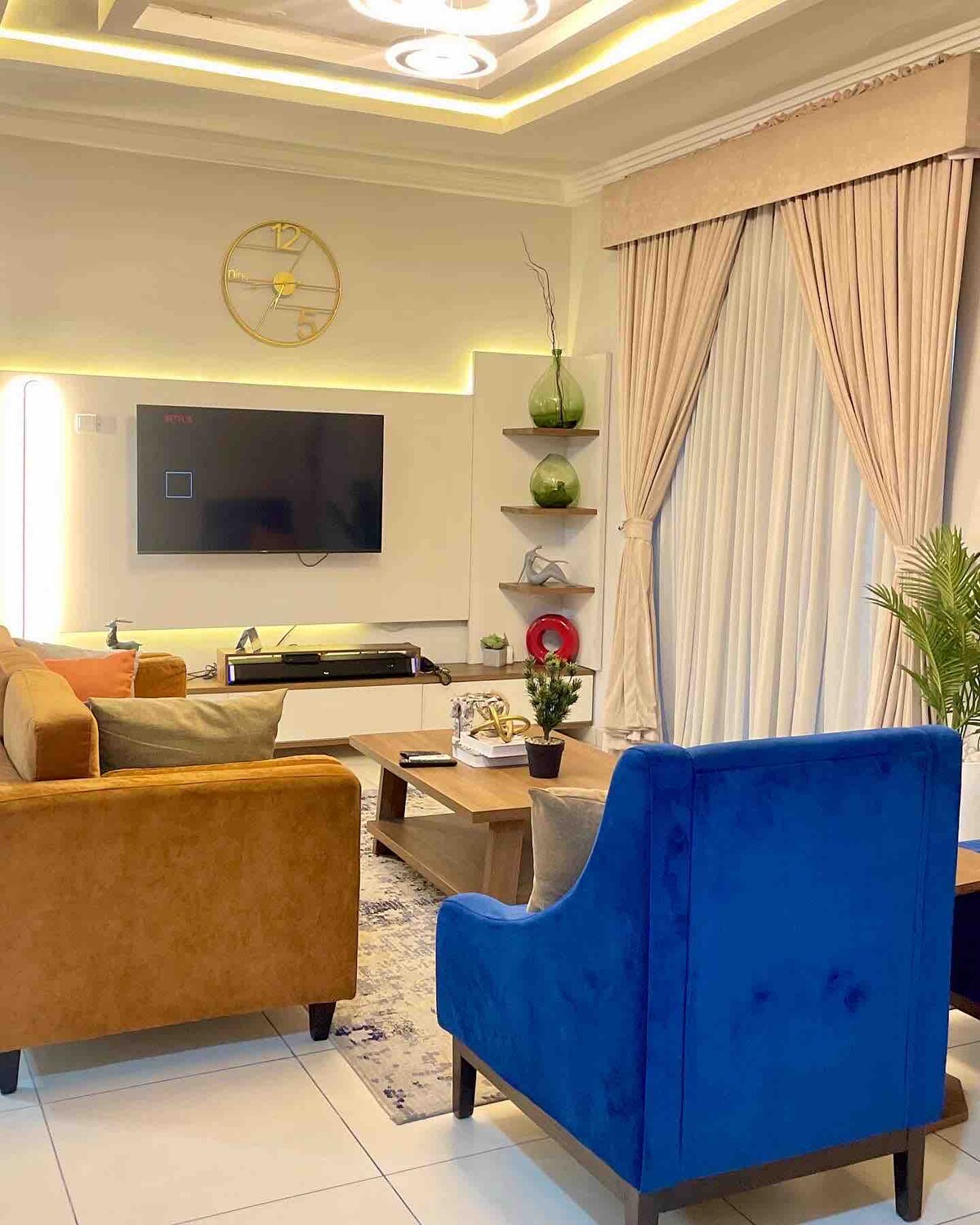 A cozy living room is showcased, featuring a large sofa in warm tones and a blue accent chair. A wall-mounted television is positioned across from the seating area, while decorative shelves with green vases enhance the space. Natural light filters through elegant curtains.