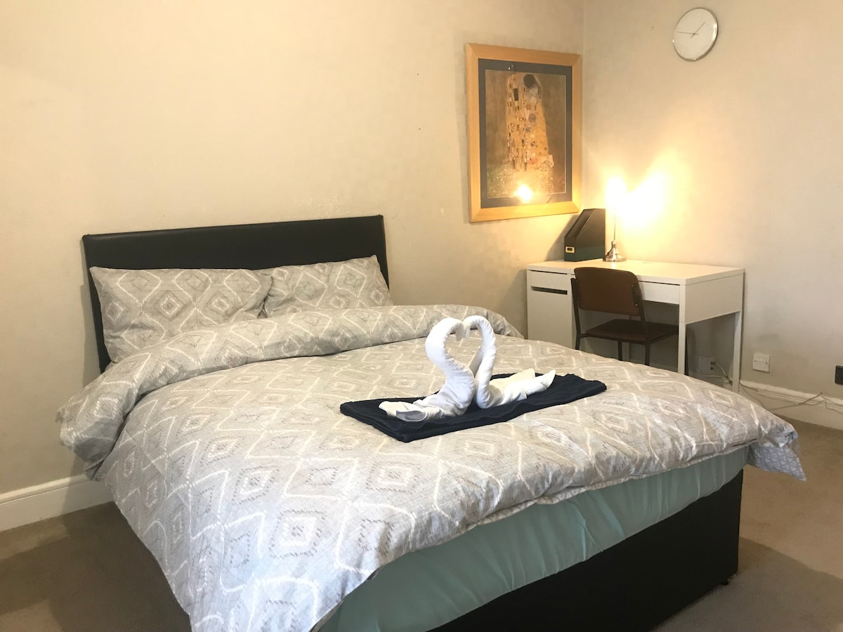 A spacious bedroom is furnished with a large bed, dressed in a patterned duvet cover. A heart-shaped towel arrangement is displayed on the bed. A study desk with a chair is positioned near the window, accompanied by a wall clock and artwork.