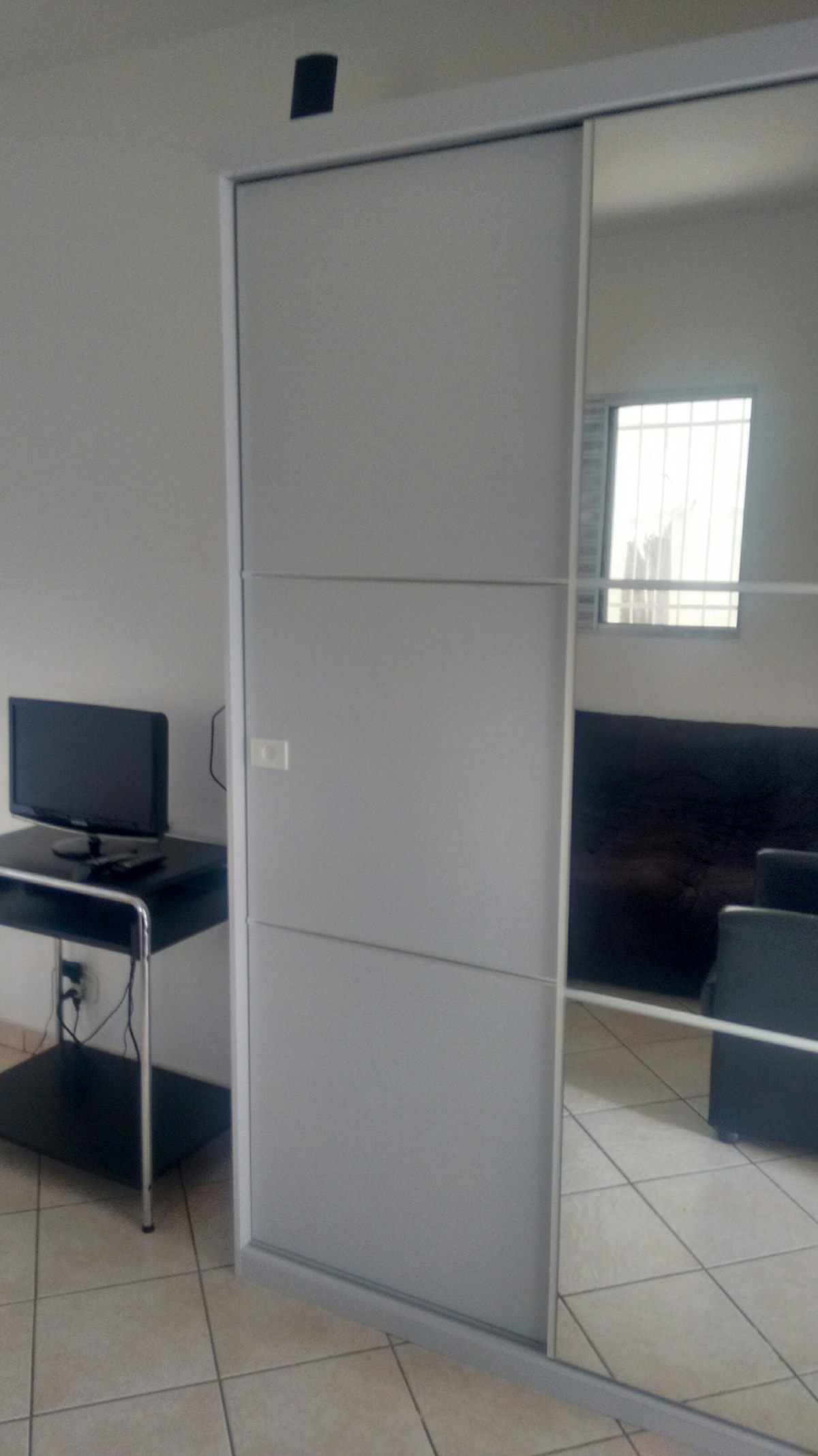 A sliding closet door, featuring a smooth white finish with mirrored panels, reflects the soft lighting of the room. A small black desk with a computer is positioned beside the closet, accompanied by a sofa in the background, featuring a neutral-colored upholstery.