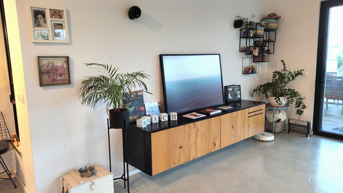 A stylish living area features a wooden sideboard with decorative items and a large flat-screen TV. Succulents and artwork adorn the walls, creating an inviting atmosphere. Large glass doors lead to an outdoor space, enhancing the connection between inside and outside.