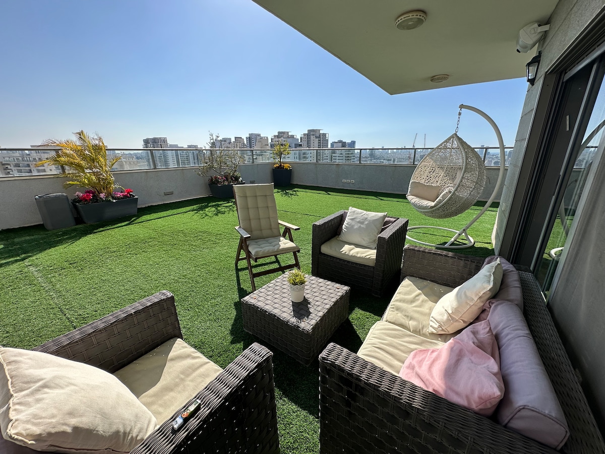 A spacious outdoor balcony is adorned with artificial grass, featuring a hanging chair and comfortable seating options. A small table is positioned at the center, surrounded by two lounge chairs, with the skyline of the city visible in the background.