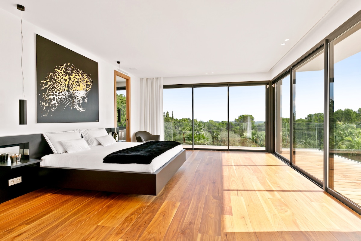 A spacious bedroom features a king size bed with a plush black throw. Large glass doors provide abundant natural light and lead to a shared balcony with garden views. A striking black and white artwork adorns one wall, complemented by warm wood flooring.