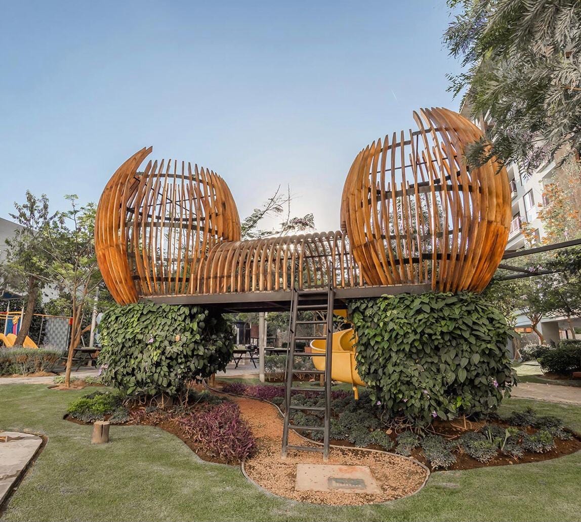 A unique wooden play structure resembles two intertwining halves, perched atop a grassy area. Surrounding foliage and colorful landscaping provide a natural backdrop, while a sturdy ladder offers access to the elevated platform. Nearby, a slide integrates seamlessly into the play area.