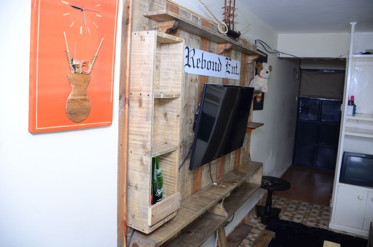 A wooden entertainment unit holds a flat-screen television with shelves for decorative items and a small bottle. A framed wall art piece featuring a quirky design is positioned above. Neutral-colored walls complement the rustic decor.